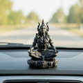Statue of a meditating figure on a car dashboard with a blurred road and trees in the background